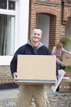 A smiling young man dressed in casual clothing, including a dark navy jacket and beige trousers, is standing outside a residential building in Knightsbridge, holding a large cardboard box with both hands. The background features a red brick house with large white-framed windows and a brick archway, situated on a residential street with cobblestone paving. Behind him, a woman is seen with light brown hair, wearing a pink sweater, and is engaged in handling packed boxes and moving supplies near the entrance of the house. Several cardboard boxes of varying sizes are stacked nearby, some sealed with packing tape, indicating a house move in progress. The overall scene captures a typical house removals process with attention to detail in the textures and colours of the cardboard, brick, and clothing, highlighting the professional service of Knightsbridge Man with Van in the local area, possibly within postcode SW1 or surrounding districts.