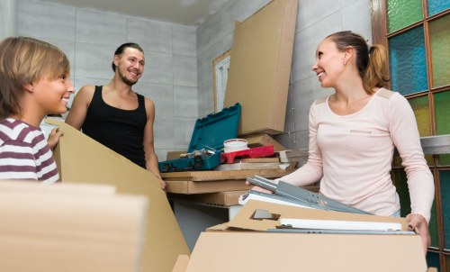 Inside a residential house, three people are engaged in a packing or moving activity, likely part of a house removal service in Knightsbridge; a woman with long brown hair tied back, wearing a light pink sweater, is smiling and holding a stack of documents or cardboard folders as she stands behind a cluttered work surface with cardboard boxes, packing tape, and tools. To her left, a young boy with blond hair, wearing a striped shirt, is looking towards her with a smile. Behind them, a woman with dark hair tied in a bun, dressed in a black tank top, is also smiling, holding a large cardboard box. The background features a wooden door frame, partially visible window with multi-coloured grids, and a plain wall, indicating a typical house interior environment. The scene suggests a friendly and organized atmosphere during a local house move, with materials and household items ready for transport, consistent with the services offered by Knightsbridge Man with Van in the area.