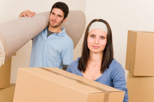 A young man and woman with light skin inside a residential property during a house move, with the man in a light blue shirt carrying a large piece of furniture wrapped in protective material over his shoulder, and the woman in a blue top holding a medium-sized cardboard box. The background features a neutral-colored wall with additional boxes stacked nearby, suggesting an active packing or unpacking process common in house removals in the Knightsbridge area. Natural lighting illuminates the scene, highlighting the textures of the cardboard boxes, the fabrics of their clothing, and the household environment. The scene is set in a typical living space, reflecting an ongoing domestic relocation activity facilitated by Knightsbridge Man with Van, known for professional removals services in the locale.