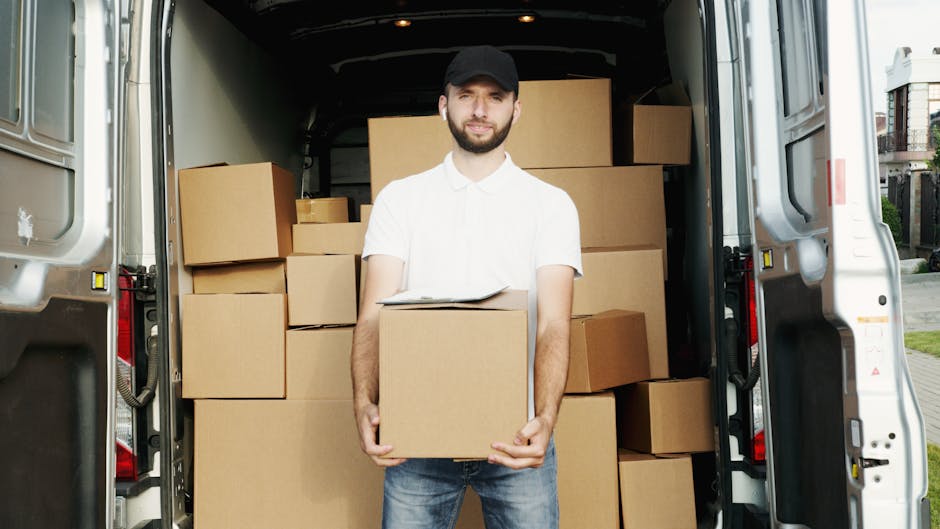 A man with a beard, wearing a black cap, white polo shirt, and jeans, stands inside the open rear of a white moving van. He is holding a medium-sized cardboard box with both hands, positioned in front