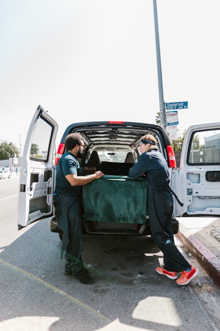 A man with a beard, wearing a black cap, white polo shirt, and jeans, stands inside the open rear of a white moving van. He is holding a medium-sized cardboard box with both hands, positioned in front