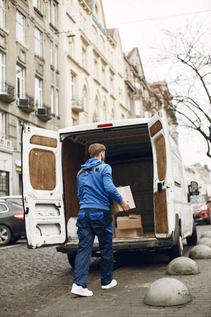 A man wearing a blue jacket and dark trousers is standing at the open rear doors of a white moving van, positioned on a cobblestone street in an urban area. Inside the van, there are several cardboard