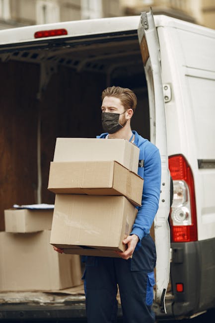 A man wearing a blue jacket and dark trousers is standing at the open rear doors of a white moving van, positioned on a cobblestone street in an urban area. Inside the van, there are several cardboard