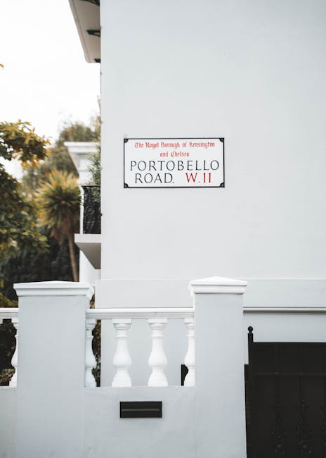 Close-up view of a house exterior showing a street sign for Portland Road, W.11, mounted on a brick wall beneath a white window frame. In the foreground, a flowering magnolia bush with large, pink and